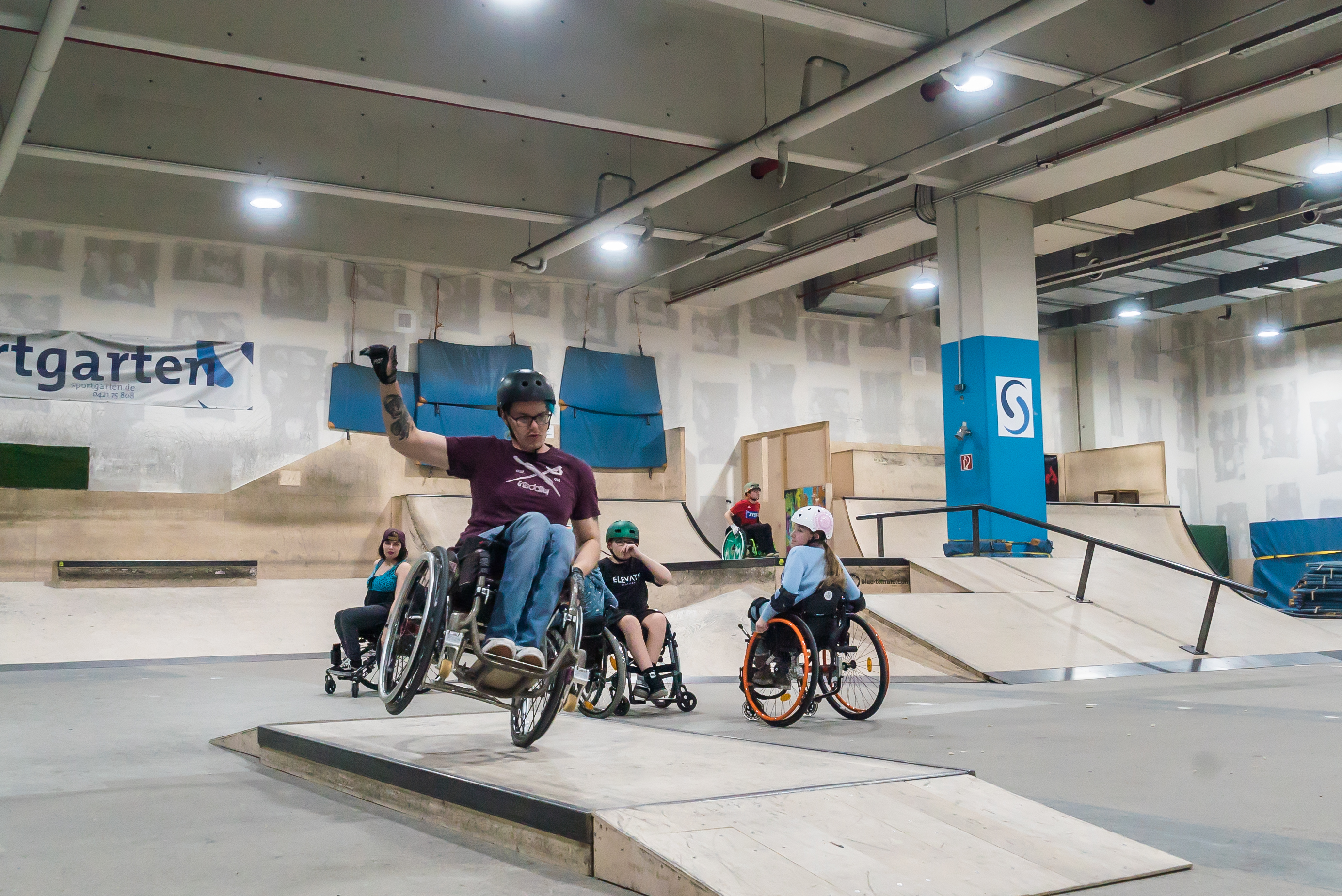F&uuml;nf Rollstuhlfahrer:innen fahren in der Skatehalle am Sportgarten &uuml;ber Rampen und Pipes.